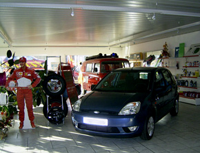 Auto in Verkaufsraum, daneben eine 'Michael Schumacher' Figur und ein Reifenstapel